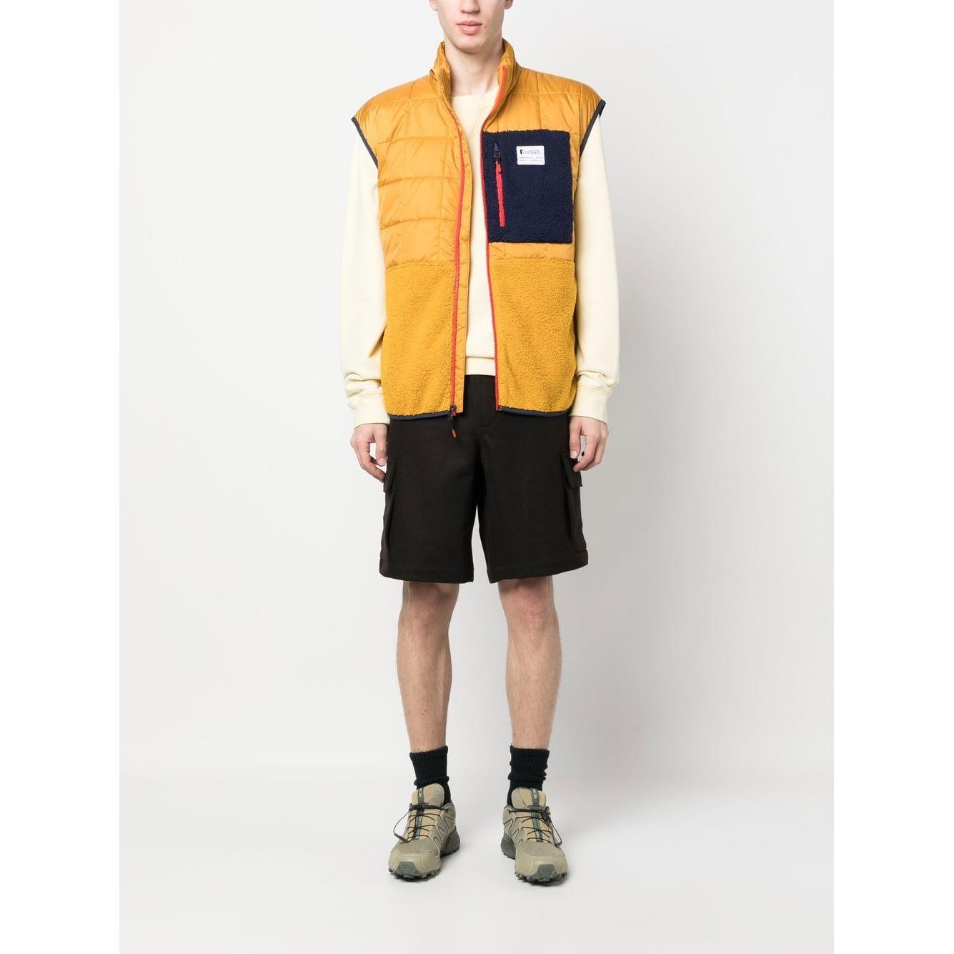 Model wearing mustard yellow COTOPAXI Trico Hybrid padded gilet with chest logo patch and front zip fastening, shown with shorts and hiking boots.