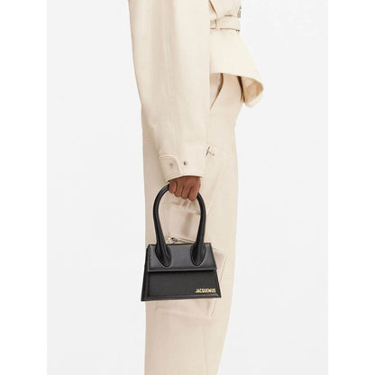 Jacquemus Le Chiquito Moyen Black Bag with gold hardware, magnetic flap, and removable shoulder strap on model's hand.